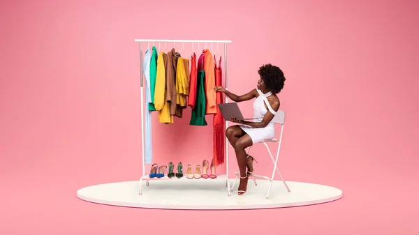 Smiling young african american woman fashion designer in white dress with laptop looks at clothes and shoes on platform in showroom on pink studio background. Fashion, style and modern device