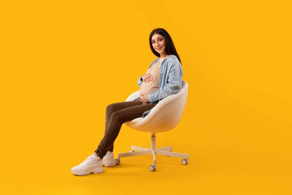 母亲的概念 兴奋的孕妇坐在黄色工作室的椅子上 温柔地抱着肚子 享受怀孕时间的孕妇 — 图库照片