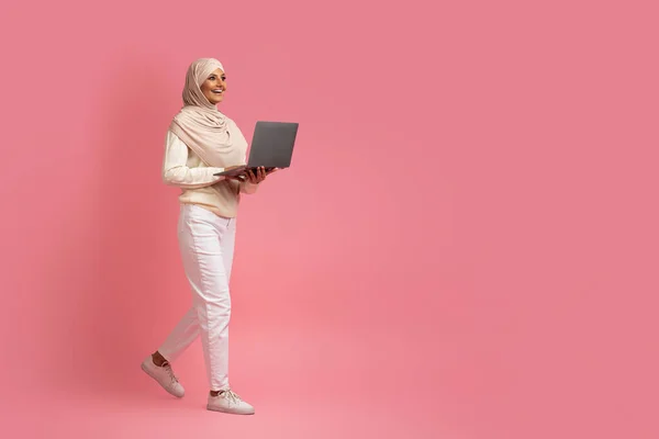 Smiling Muslim Woman In Hijab Walking With Laptop Over Pink Studio Background, Full Length Shot Of Happy Islamic Female In Headscarf Using Computer For Remote Work Or Online Shopping, Copy Space