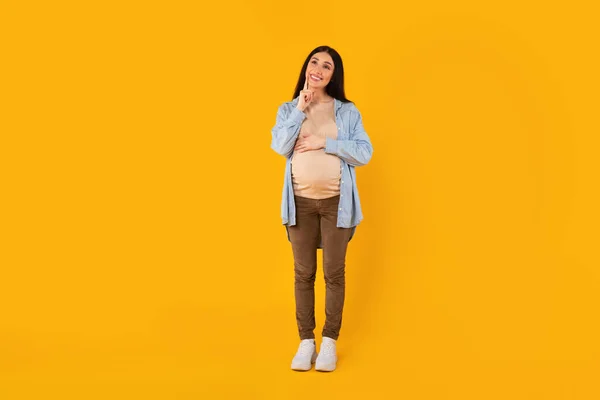 快乐的沉思的孕妇摸着肚子和下巴 抬起头思考着问题 梦想着宝宝的性别 未来的分娩 站在黄色工作室的背景上 — 图库照片