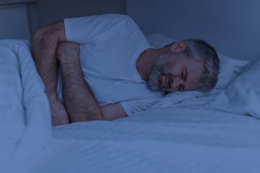 Mutsuz, orta yaşlı, kır saçlı, sakallı bir adam evde yatarken acı çekiyor, karnına dokunuyor, karın ağrıları çekiyor, geceleri karın ağrısı çekiyor, ev içi, fotokopi odası.