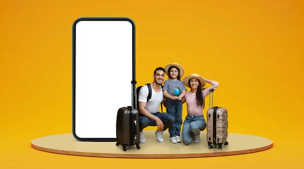 Glad millennial arab family with small daughter enjoy vacation with suitcases, globe on stage on orange studio background with huge smartphone with empty screen. App for trip, travel