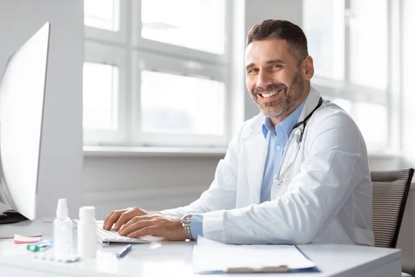 病院でコンピュータを使用して白衣の幸せな男性医師医学の専門家 患者の電子ファイルの医療フォームをチェック オンライン仮想相談を持っています カメラを見て笑顔 — ストック写真