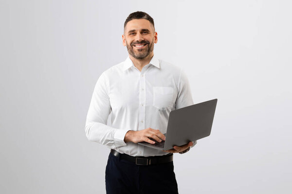 Portrait of cheerful middle aged businessman with laptop computer in hands smiling at camera, standing on light grey background, chatting with clients online