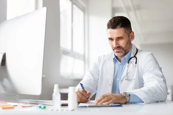 严重的男性医生使用计算机和写笔记在医学期刊坐在诊所的工作场所 穿着白衣在办公室工作的专职内科医生 — 图库照片
