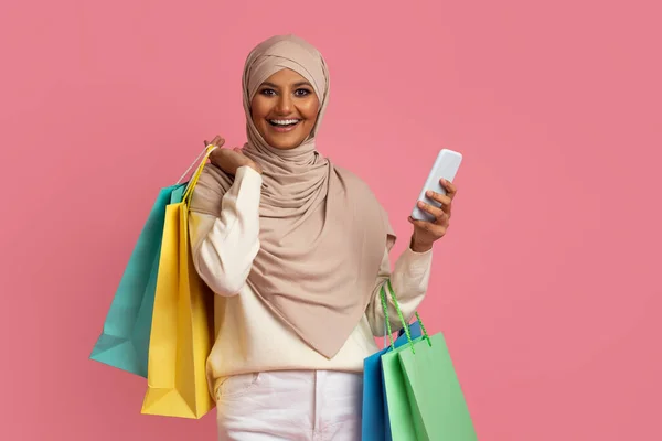 购物应用程序 在Hijab手持智能手机和购物袋 快乐的中东购物狂女士使用移动应用程序 站在粉色背景之上 复制空间中微笑的穆斯林妇女 — 图库照片