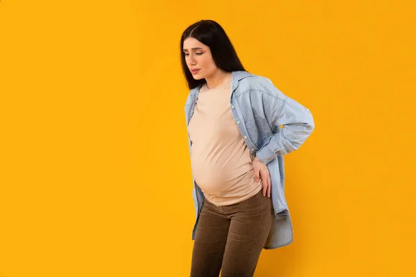 卫生保健概念 年轻的孕妇遭受情人背痛 摩擦发炎的部位以减轻疼痛 站在孤立的黄色背景上 复制空间 — 图库照片