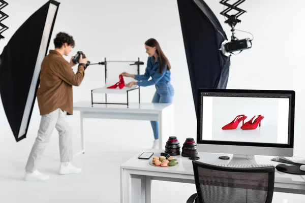 Male photographer and female art director taking photos of red shoes, working together in modern photostudio, making content photoshoot, focus on monitor with picture