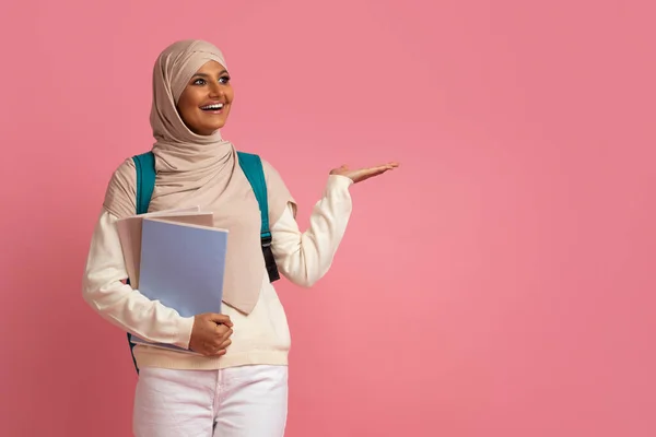 Smiling Arab Woman In Hijab Pointing Aside At Copy Space With Her Palm, Happy Muslim Female Wearing Headscarf And Backpack Showing Free Place For Advertisement While Standing On Pink Background