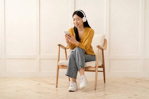 白い壁の横にあるアームチェアに座って 自宅でスマートフォンやワイヤレスヘッドフォンを使用して 女性は音楽に耳を傾け ビデオコンテンツを見て コピースペース — ストック写真