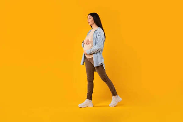 カジュアルな服装の若い妊婦 歩くと黄色のスタジオの背景に彼女の腹に触れると フル長さのショット フリースペース 幸せな妊娠時間 — ストック写真