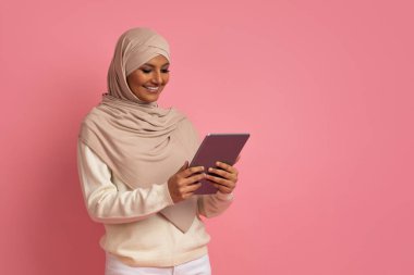 tesettürde gülümseyen Müslüman kadın dijital tablet kullanıyor, internetten alışveriş yapıyor veya sosyal ağlara göz atıyor, başörtüsü takan Arap kadın modern teknolojilerin tadını çıkarıyor, Pembe Stüdyo Arkaplanının başında duruyor