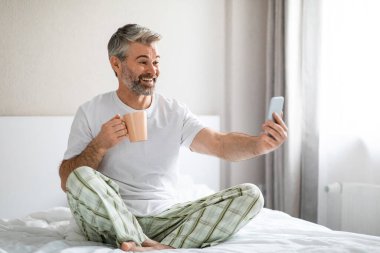 Duygusal olarak mutlu, gülümseyen, gri saçlı, pijama giyen, elinde kahve fincanı ve akıllı telefon tutan, uyandıktan sonra sevgilisiyle görüntülü konuşma yapan, uzay fotokopisi çeken orta yaşlı bir adam.