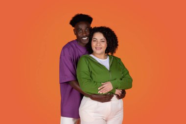 Kırmızı arka planda kucaklaşan ve gülümseyen neşeli, genç, sevgi dolu melez bir çiftin stüdyo portresi. Afrikalı Amerikalı bir adam İspanyol tombul bir kadını kucaklıyor.