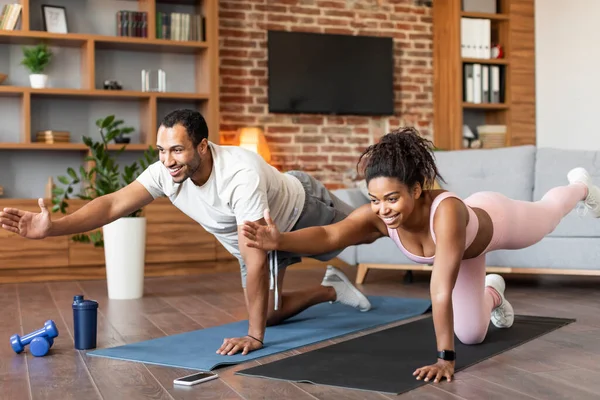 Sport lifestyle, family body care at home. Smiling young black guy and female in sportswear doing leg exercises, make plank together on floor in living room interior. Workout, fitness and relationship