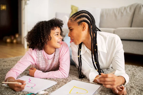 Funny African American Mommy And Her Preteen Daughter Having Fun Sticking Out Tongues Drawing Together Lying On Floor At Home. Family Bonding On Weekend. Hobby And Leisure Concept