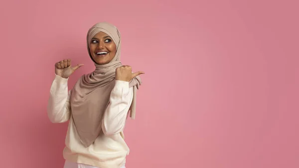看那里 身穿头巾的快乐的穆斯林妇女竖起大拇指 指着她身后的复制空间 兴奋的阿拉伯女士站在粉红的工作室背景 全景之上 示意着可以登广告的地方 — 图库照片