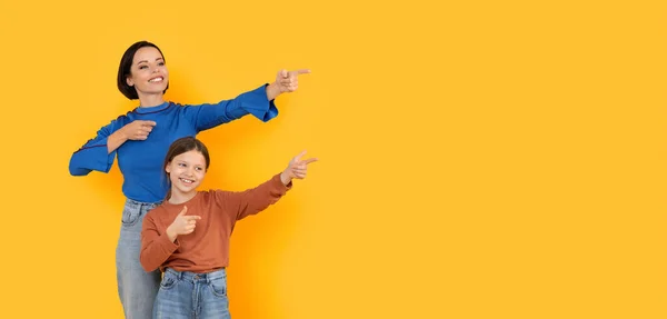 看看这个快乐的妈妈和小女儿带着两个小指 快乐的妈妈和可爱的女儿 在复制的空间里指指点点站在黄色背景下的广告的免费位置 — 图库照片