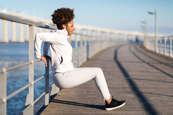 stock image Young black woman in sportswear squatting outdoors, using handrail as support, athletic african american female doing sit-up exercise while practicing sports outside at wooden pier near sea