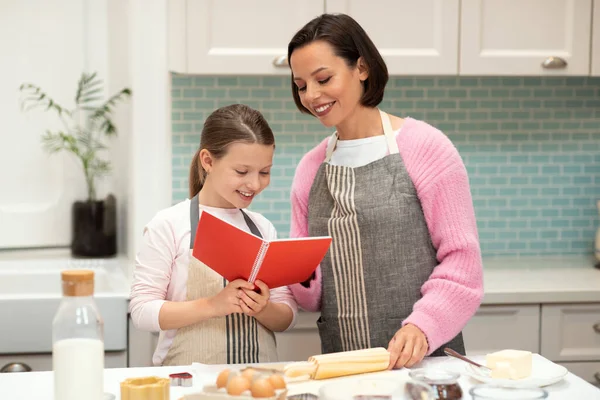 Glad caucasian young mom and small daughter in aprons make cookie dough, look at recipe book in kitchen interior. Cooking sweets together, lesson of homemade food, hobby at home