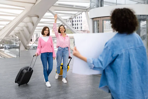 Two women waving to their female friend meeting them with name board at airport after arrival, happy ladies waling with suitcases and smiling, excited to see each other after trip abroad
