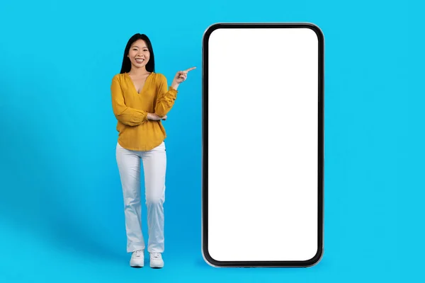 快乐快乐的笑着漂亮的年轻韩国女人在网上展示了巨大的智能手机与白色空白屏幕模型 隔离在蓝色的工作室背景 复制空间 — 图库照片