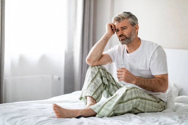 Unhappy middle aged man in pajamas suffering from morning headache after sleeping in bedroom, sitting on bed after waking up, touching his head, holding glass of water, looking at copy space