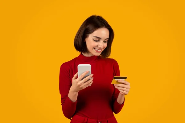 笑着穿红色衣服的漂亮年轻女人 手里拿着现代智能手机和信用卡 背景是橙色的工作室 为商品和服务付费 在网上购物 复制空间 — 图库照片