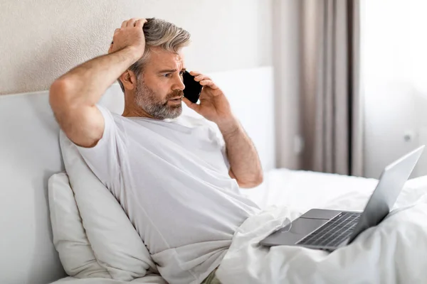 Troubled stressed upset handsome grey-haired middle aged man sitting in bed, working from home, using laptop, have phone call, wearing pajamas, bedroom interior, copy space, side view