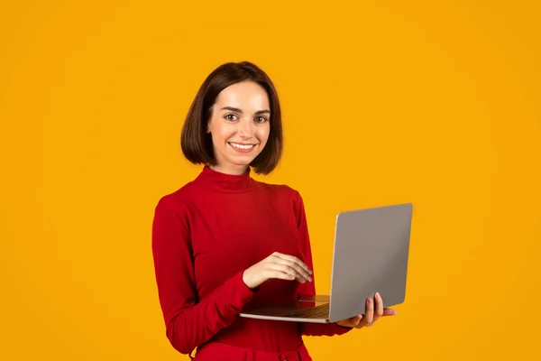 幸せな魅力的なブルネット若い女性で赤デジタル遊牧民で現代のPcラップトップを使用してオレンジ色のスタジオの背景に コンピュータのラップトップを入力し カメラで笑顔 楽しい素敵なデバイス — ストック写真