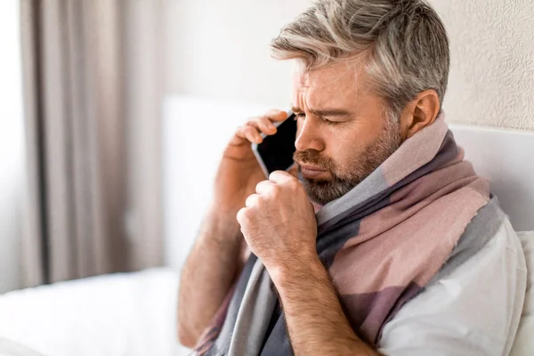Sad sick middle aged grey-haired man with warm scarf around his neck sitting in bed at home, talking the phone to his employer while asking about the sick leave, calling doctor, coughing, copy space