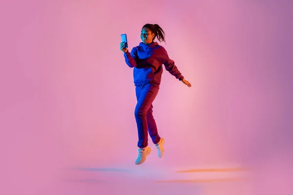 全身の長さショットの若いです黒女性でスポーツウェアジャンプと上の携帯電話を使用してピンクネオンライトカラー背景 フリーコピースペース — ストック写真