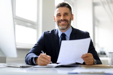 Takım elbiseli mutlu orta yaşlı işadamı iş yerindeki belgelerle çalışıyor ve kameraya gülümsüyor, şirketin CEO 'su mali raporları tutuyor, boş yer var.