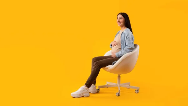 妊娠時間 興奮した妊婦の椅子に座って 彼女の腹を愛撫し 脇に黄色の背景に無料のスペースを見て パノラマ 期待の女性優しく受け入れるおなかと笑顔 — ストック写真