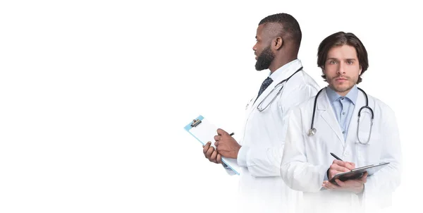 手にクリップボードと制服を着た2人の男性医師との複合画像 白い背景に隔離された立っている医療コートを身に着けている多民族医療従事者 コピースペースとパノラマ — ストック写真