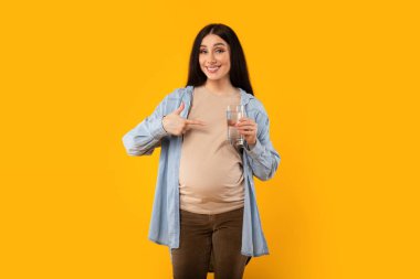 Mutlu hamile kadın bir bardak su tutuyor ve onu işaret ediyor sarı stüdyo arka planındaki kameraya gülümsüyor. Aqua dengesi, sağlık, diyet ve yararlı beslenme