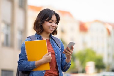 Gülümseyen Arap Koleji öğrencisi akıllı telefon kullanıyor ve derslerden sonra dışarıda dinlenirken çalışma kitaplarını tutuyor neşeli Orta Doğulu kadın cep telefonuyla mesaj atıyor ya da yeni uygulamalara bakıyor, Boşluğu kopyalıyor.