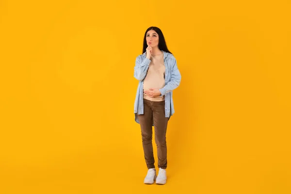 漂亮的女人在等着孩子 摸着怀孕的肚子 手拉着下巴 在黄色的背景下思考问题 全身而退 — 图库照片