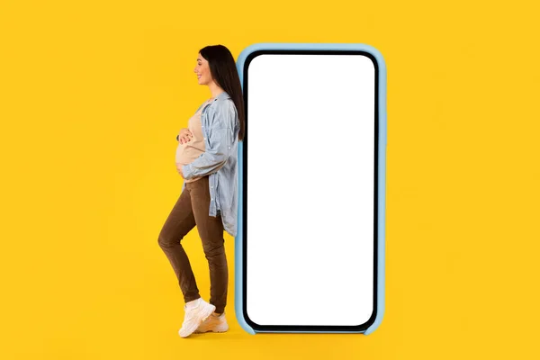 妊娠アプリのためのモバイルモックアップ 妊娠中の女性は 空白の画面で巨大な携帯電話で傾いて 彼女の腹を受け入れます 広告オンラインオファーやウェブサイト コラージュ — ストック写真