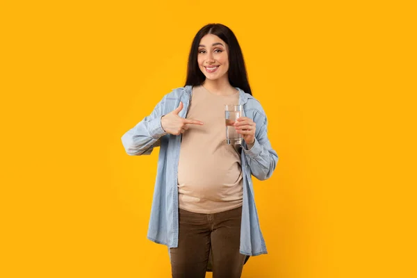 快乐的孕妇拿着一杯水指着它 在黄色的工作室背景上对着相机微笑 水产平衡 饮食和有益营养 — 图库照片