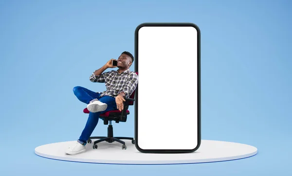 Laughing young african american guy call by smartphone, talk, sit in armchair with huge smartphone with blank screen on platform on blue studio background. App, website communication, good news