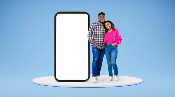 快乐的年轻的非洲裔美国夫妇在随意的拥抱下 呆在大手机旁边 蓝色工作室背景 屏幕空荡荡 人际关系 应用程序和设备网站 — 图库照片