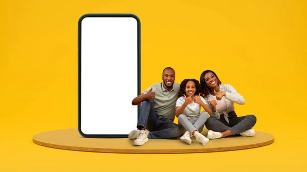 stock image Smiling african american millennial family with teen daughter show thumbs up gesture, approving or recommend ad and offer on huge phone with empty screen on yellow studio background