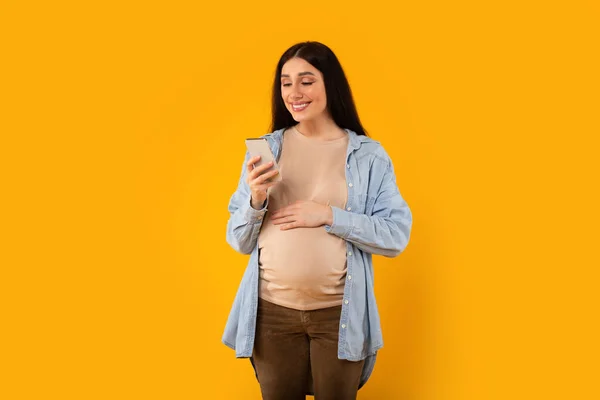 黄色の背景の上に携帯電話でチャットしながら 陽気な若い妊婦立ってポーズ 腹に触れます スマートフォンや笑顔を使用して — ストック写真