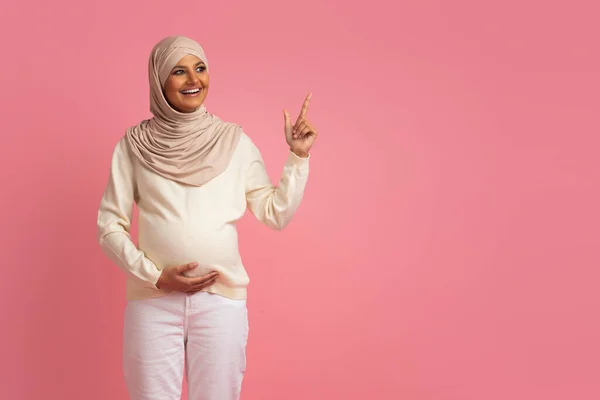 中东孕妇头戴头巾 站在粉红工作室的背景上摸着肚子 指着复制的空间 表示可以免费登广告 — 图库照片