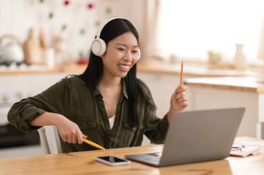 Rahat genç Asyalı kadın evde çalışırken mola veriyor, mutfak masasında oturuyor, elinde kalemler, kablosuz stereo kulaklıklar ve modern dizüstü bilgisayar kullanıyor, müzik dinliyor, boşluk kopyalıyor.