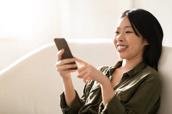 長い絹のような髪を持つ幸せな陽気なかわいい魅力的な中国の女性は スマートフォンでソファに座って ブログを読んで ソーシャルメディアでスクロールし 友人とチャット 自宅のインテリア クローズアップ コピースペース — ストック写真