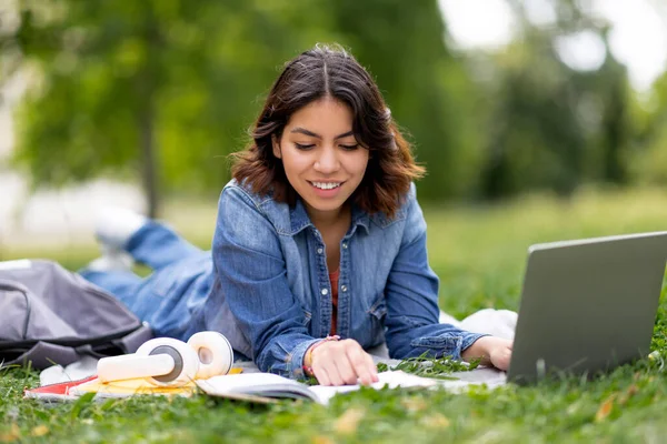 中东快乐的年轻女性阅读书籍 躺在公园草坪上躺着用电脑学习的户外写真 — 图库照片
