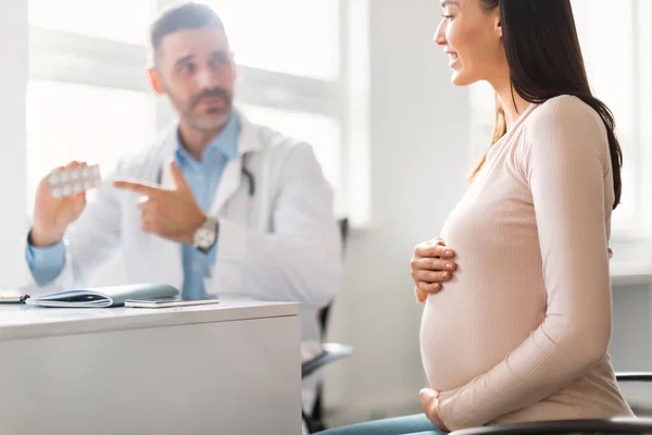 Pregnancy and medicine concept. Professional male gynecologist giving pills to young pregnant lady during appointment at hospital, future mom visiting doctor, selective focus