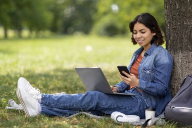 Modern Teknolojiler. Zeki telefonlu ve laptoplu gülümseyen Arap kadın dışarıda dinleniyor, şehir parkındaki ağacın altında oturan mutlu Orta Doğulu kız öğrenci cep telefonundan mesaj atıyor,
